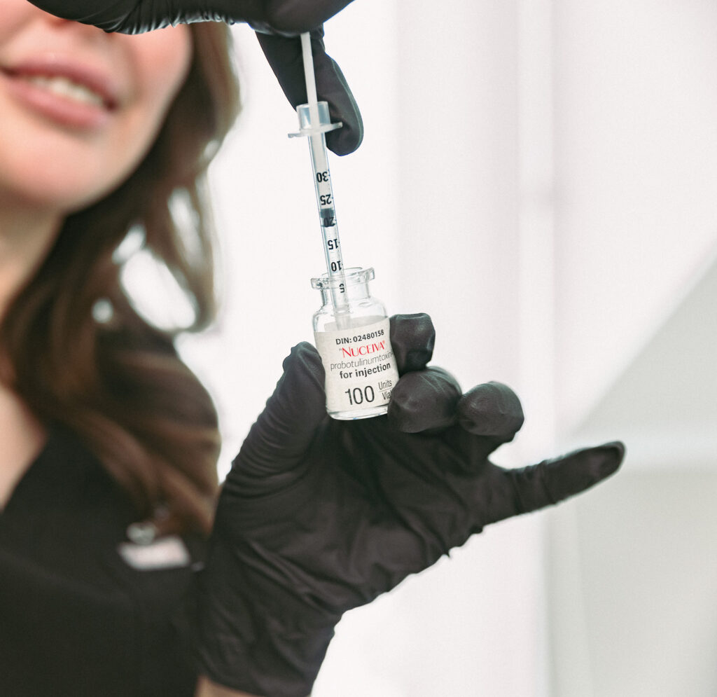 A provider wearing black gloves and holding a syringe smiles as she prepares crow's feet Botox shots in Burlington.