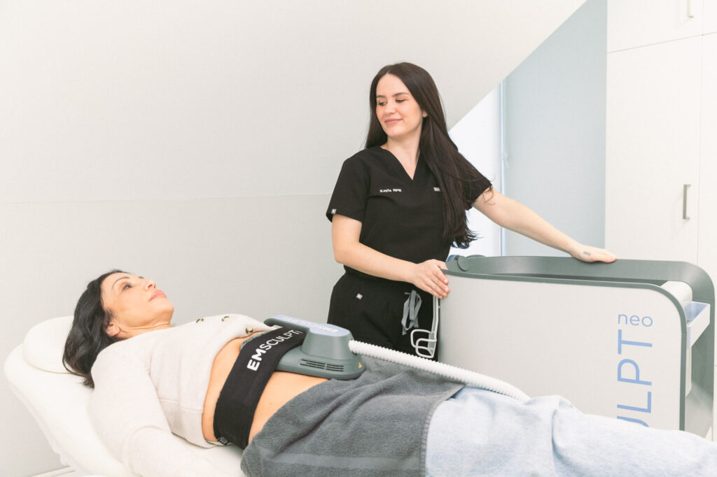 A provider in black scrubs smiles at a reclining patient as they receive Emsculpt NEO near Hamilton.